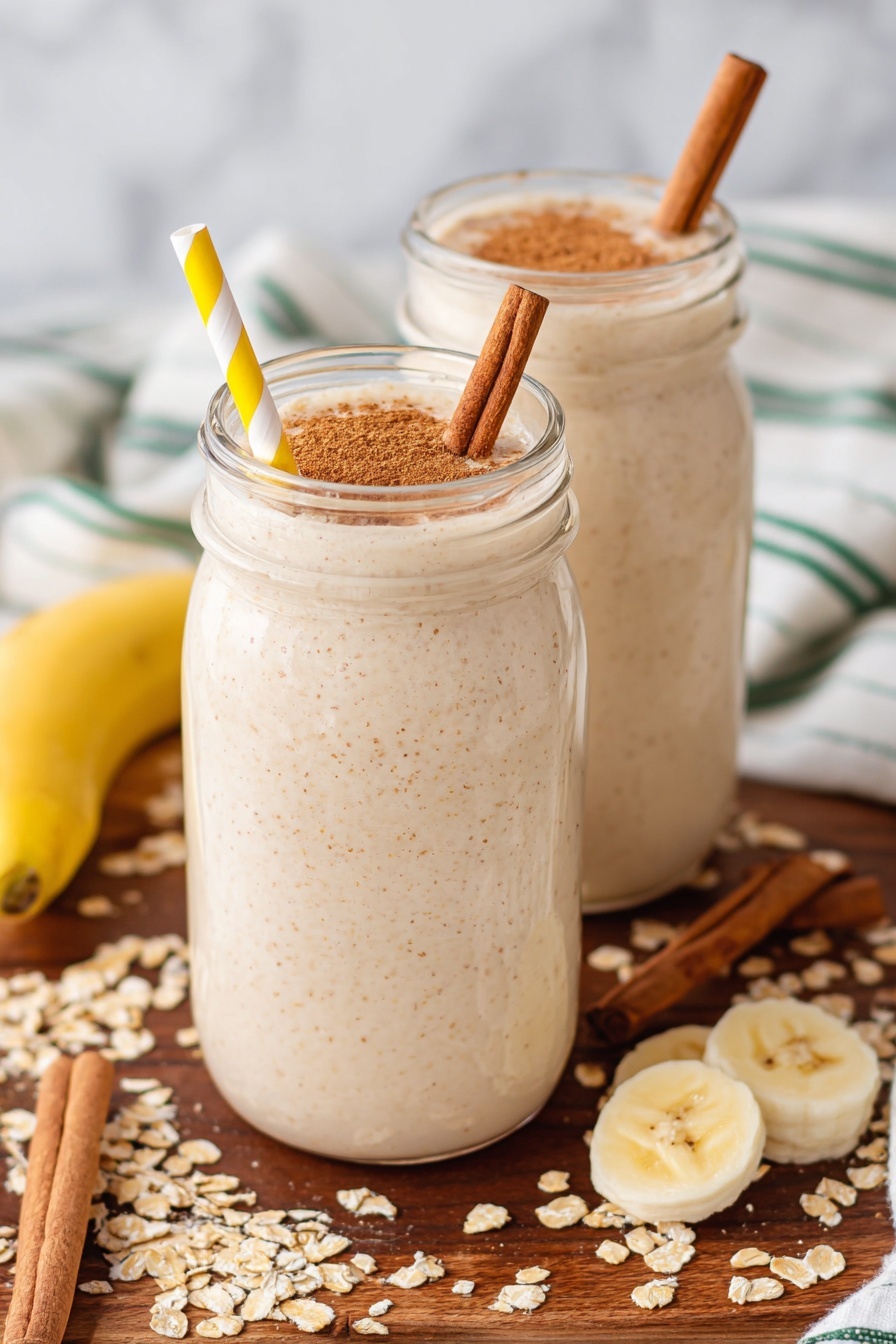 Two tall clear glass jars are filled with a creamy beige smoothie with tiny brown specks. On top of each smoothie is a layer of light brown powder sprinkled evenly and a single cinnamon stick standing diagonally inside. Each jar has a yellow and white striped paper straw. The jars sit on a wooden surface with scattered oat flakes, cinnamon sticks, and banana slices around them. In the background, a white cloth with green stripes lays on the surface. The surface is a white marbled texture. photo taken with an iphone --ar 2:3 --v 7