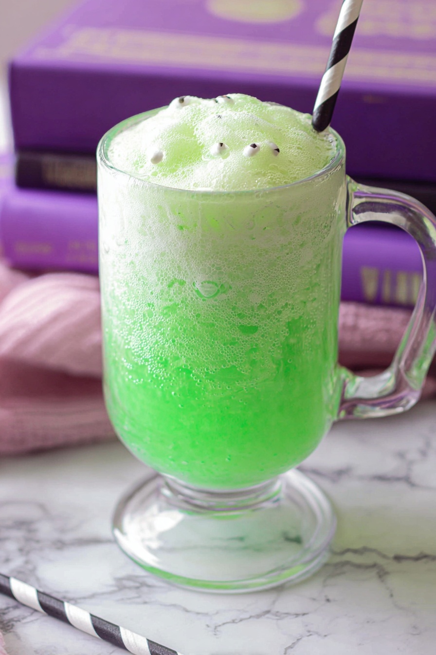 The image shows a clear glass mug filled with a bright green fizzy drink as the bottom layer. On top is a foamy pale green layer with a light, bubbly texture and small white round sprinkles with black dots scattered on it. A striped black and white paper straw is placed inside the mug on the right side. The mug sits on a white marbled surface with a purple cloth and a pink book under it. photo taken with an iphone --ar 2:3 --v 7