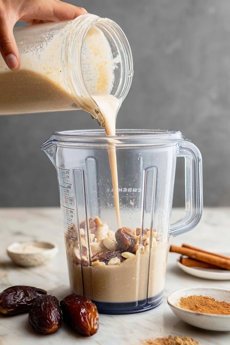 A clear blender jar filled with mixed creamy beige and light brown ingredients like nuts and dates, with a creamy light beige liquid being poured from a glass bottle by a woman's hand into the jar from above; the jar sits on a white marbled surface, and there is a white plate in the background holding cinnamon sticks, a small bowl of cinnamon powder, and some dates. photo taken with an iphone --ar 2:3 --v 7