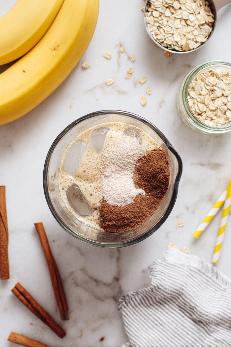 A clear glass blender is shown from above, filled with ice cubes and topped with layers of light tan powder and dark brown powder, resting on a white marbled surface. To the left, two yellow bananas and three cinnamon sticks lie close by, while a metal scoop filled with raw oats is slightly scattered nearby. On the right side, two empty clear glass jars and two yellow-and-white striped straws are visible, along with a white and gray striped cloth at the bottom right corner. Photo taken with an iphone --ar 2:3 --v 7